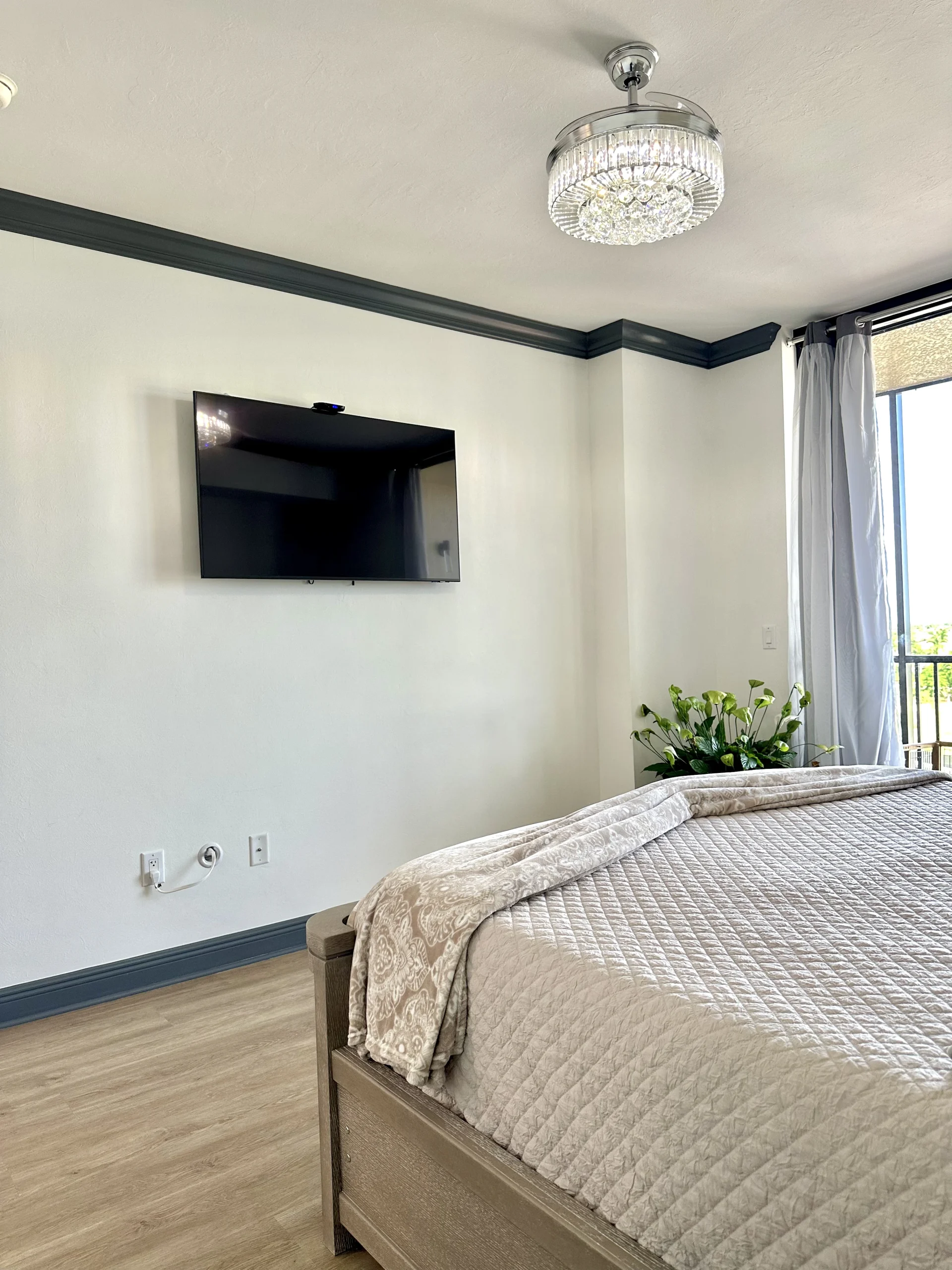 Remodeled bedroom with dark gray crown molding, a wall mounted TV and a crystal ceiling fan light in an Alliance Construction remodel in Southwest Florida.
