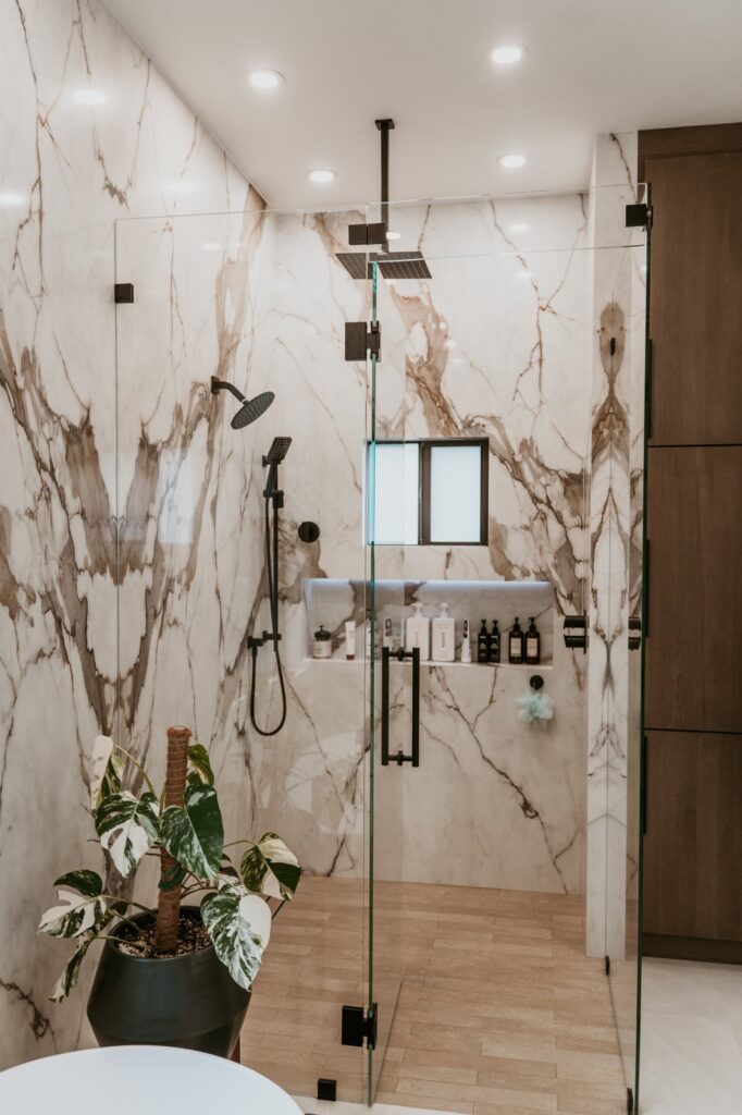 Frameless glass walk-in shower, marble walls, black rain head, niche shelf.