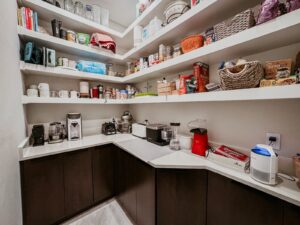 Walk-in pantry with white L-shaped shelving, coffee station, dark lowers, white counter.