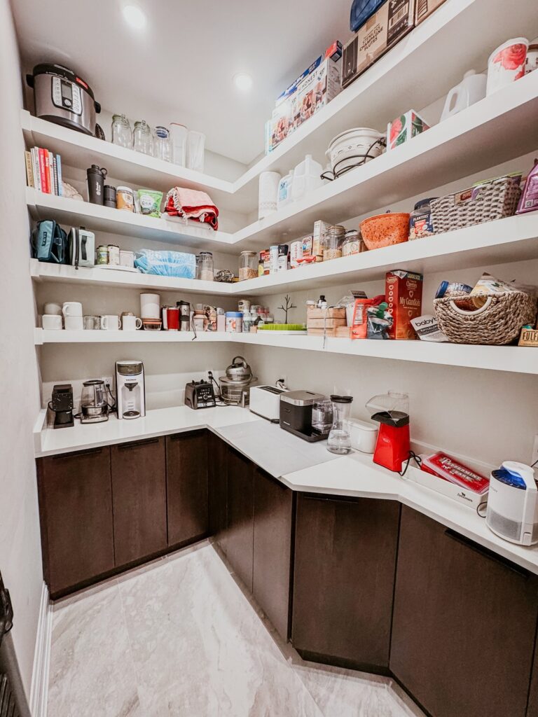 Walk-in pantry with white floating shelves, dark base cabinets and counter with small appliances.