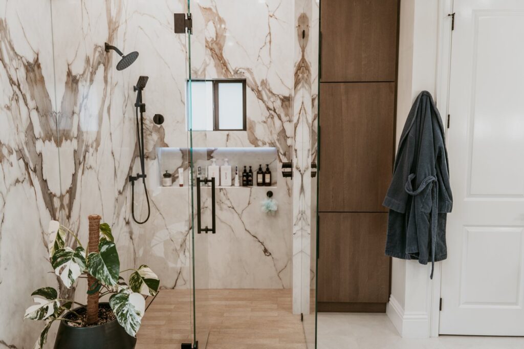 Marble walk-in shower with black rainfall, niche, plant, wood linen cabinet.