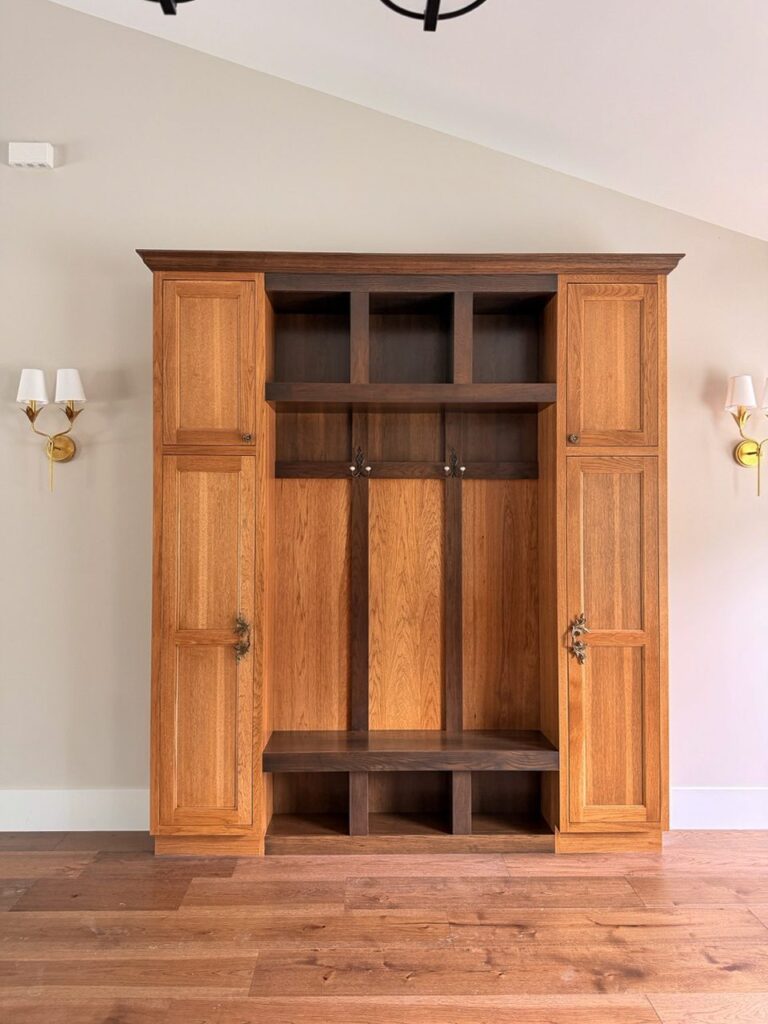 Built-in oak drop zone with tall cabinets, hooks, cubbies and a bench between brass wall sconces