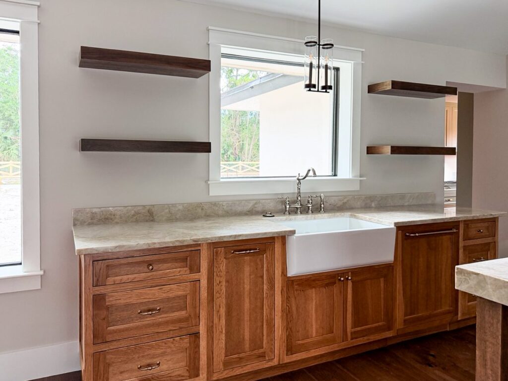 Stained oak sink run with a white farmhouse sink and bridge faucet under a picture window, framed by dark floating shelves