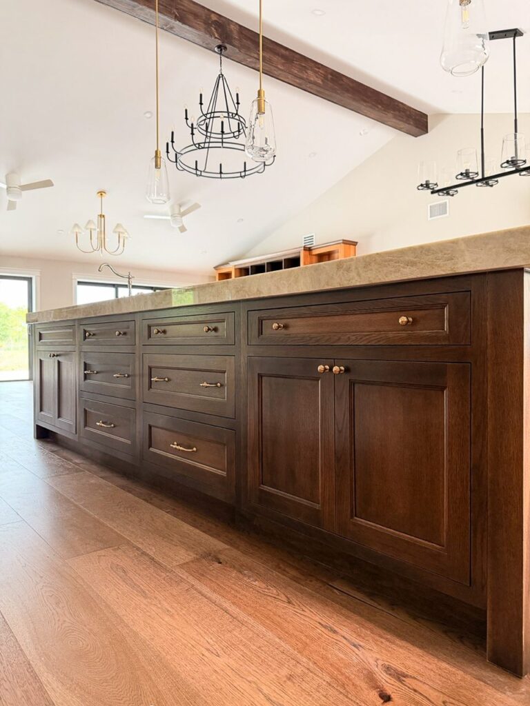 Long dark stained kitchen island drawer bank with brass pulls under a beamed vaulted ceiling with glass pendants