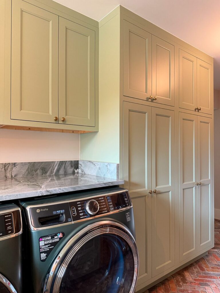 Full height sage green storage towers and upper cabinets beside a front load washer and stone counter