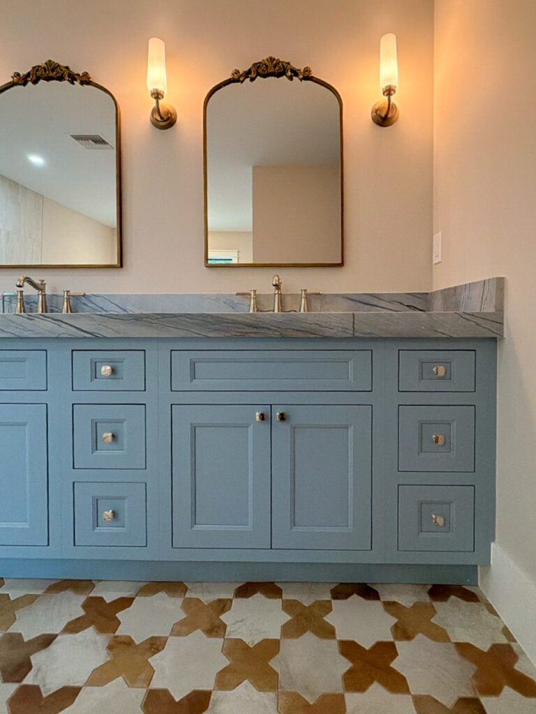 Primary bath with a blue painted double vanity, blue veined stone top, arched brass mirrors and a patterned tile floor