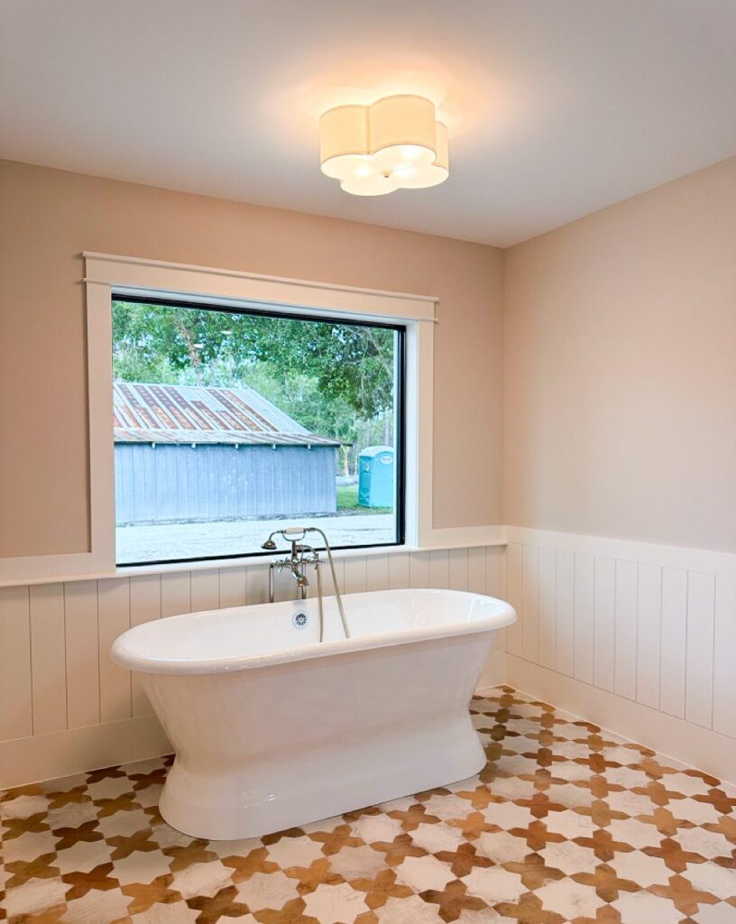 Freestanding pedestal tub under a picture window with white V-groove wainscot and a patterned tile floor
