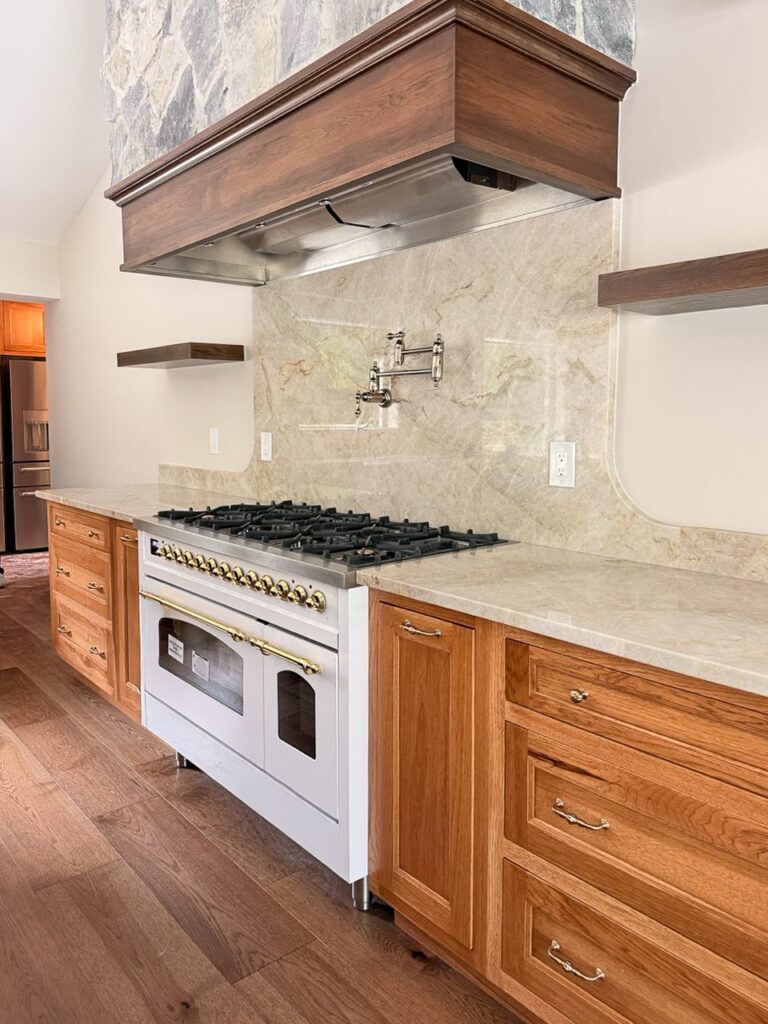White and brass range under a wood hood with a full height stone slab backsplash, pot filler and stained oak drawer stacks