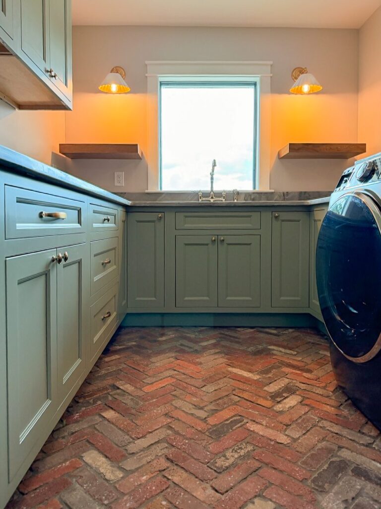 Sage green laundry room cabinetry with a herringbone brick floor, stone counters and brass sconces flanking the window