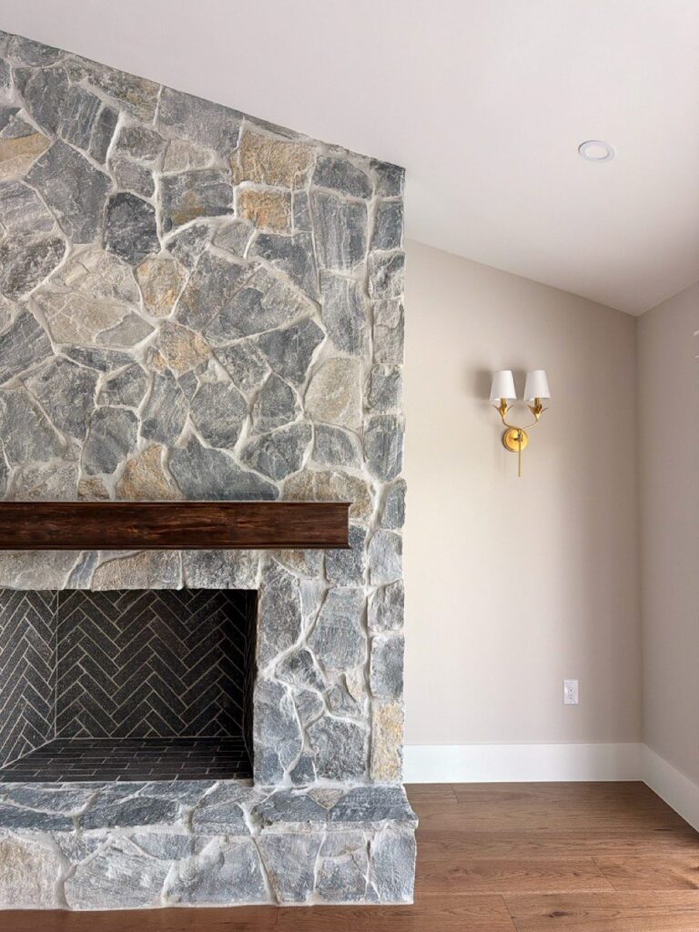 Finished stacked stone fireplace with herringbone firebox, wood mantel and brass sconce beside a sliding glass door